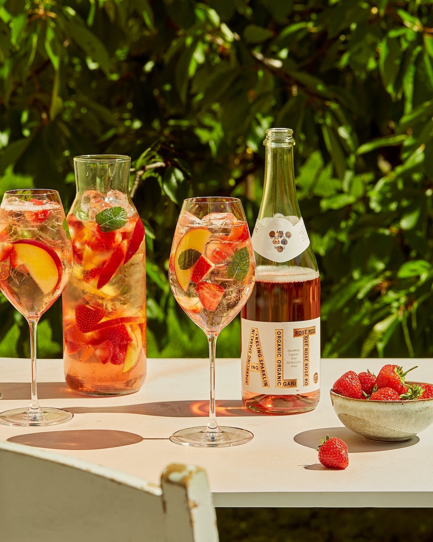 Bilde av ALT. Sparkling Rosé ute på et solfylt hagebord, med en mugge bestående av fersken, mynte, jordbær, ALT. og isbiter.